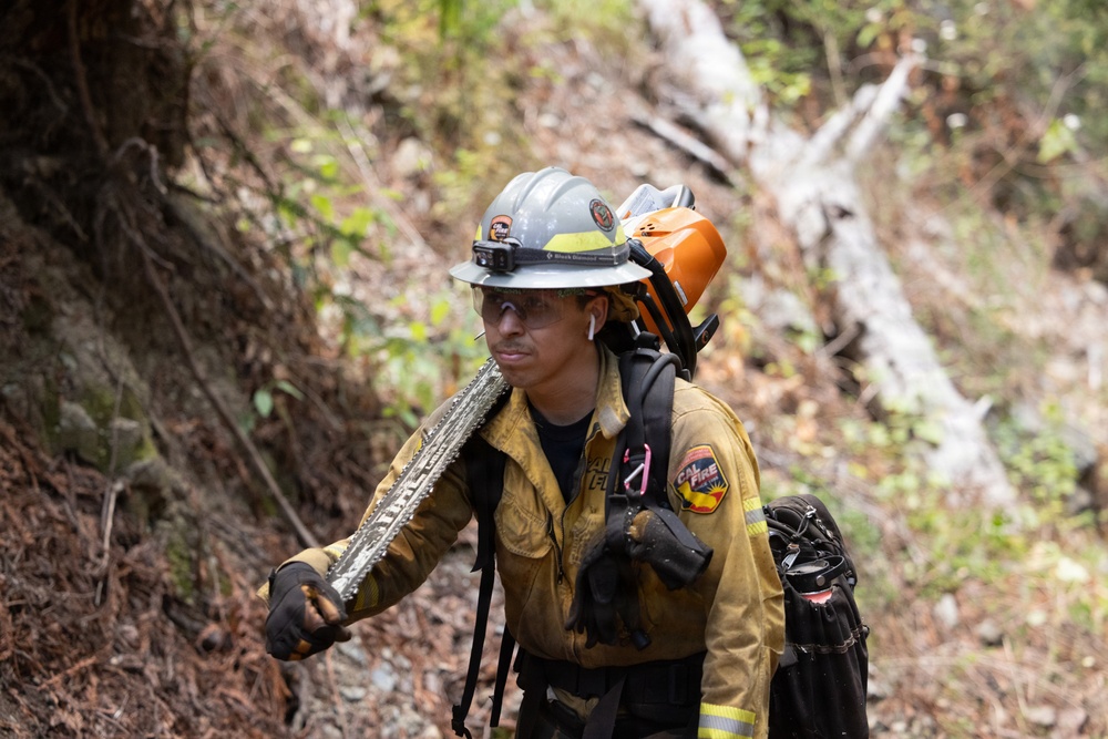 A CAL FIRE firefighter is navigating through a wooded, hilly terrain while carrying firefighting equipment. The firefighter is equipped with a chainsaw bar, hard hat with headlamp, and is carrying what appears to be a chainsaw or power tool on their back. The setting suggests active wildland firefighting or fire line preparation operations.