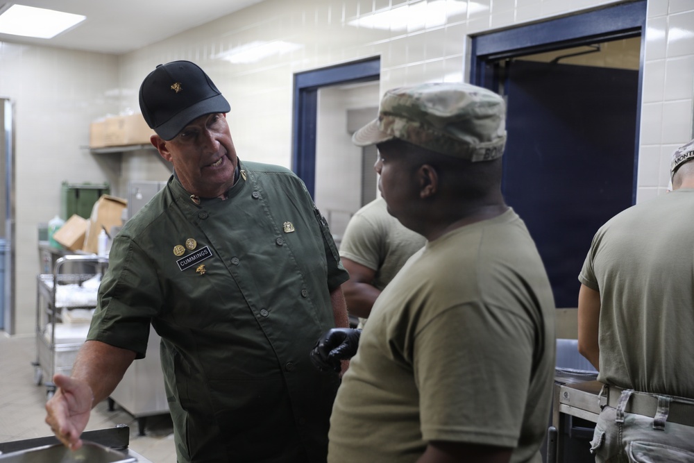 The image shows a military kitchen environment where a senior figure wearing a green uniform jacket with a name tag reading 'CUMMINGS' and multiple pins is conversing with a soldier in Army Combat Uniform (ACU). The setting appears to be a military dining facility or mess hall, with stainless steel equipment and tiled walls visible in the background. A third uniformed individual is partially visible on the right side of the frame.
