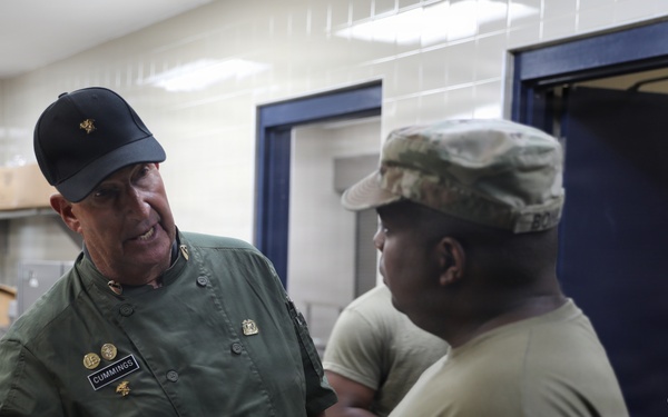 The image shows a military kitchen environment where a senior figure wearing a green uniform jacket with a name tag reading 'CUMMINGS' and multiple pins is conversing with a soldier in Army Combat Uniform (ACU). The setting appears to be a military dining facility or mess hall, with stainless steel equipment and tiled walls visible in the background. A third uniformed individual is partially visible on the right side of the frame.