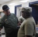 The image shows a military kitchen environment where a senior figure wearing a green uniform jacket with a name tag reading 'CUMMINGS' and multiple pins is conversing with a soldier in Army Combat Uniform (ACU). The setting appears to be a military dining facility or mess hall, with stainless steel equipment and tiled walls visible in the background. A third uniformed individual is partially visible on the right side of the frame.