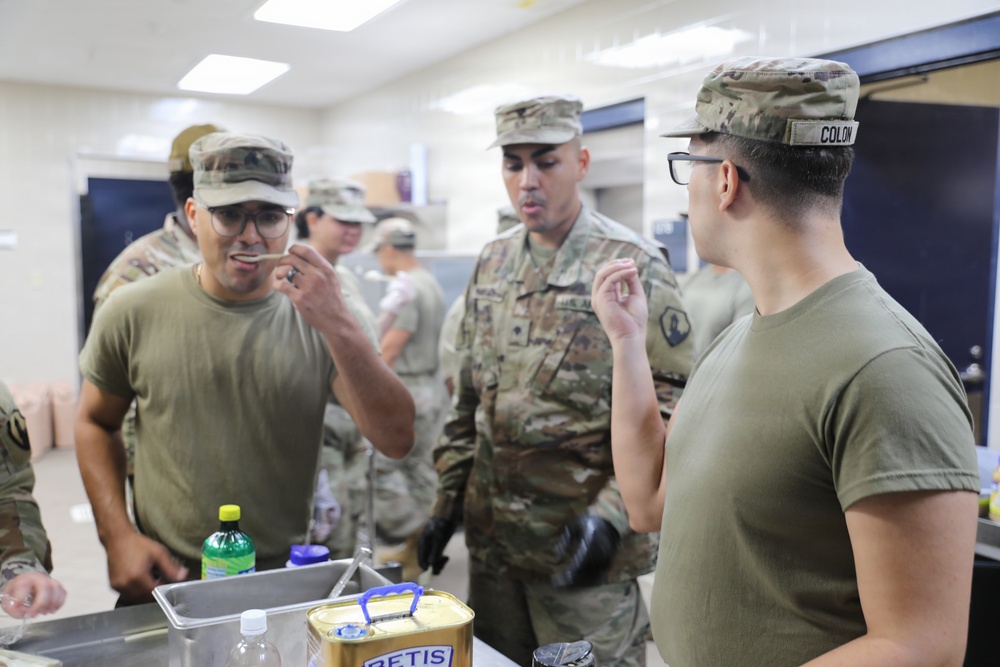 Military personnel in U.S. Army OCP (Operational Camouflage Pattern) uniforms are gathered in what appears to be a military kitchen or dining facility. Several soldiers are tasting or sampling food, with one soldier wearing a name tape reading 'COLON' visible on the right. The setting suggests a food preparation, quality control, or culinary training activity.