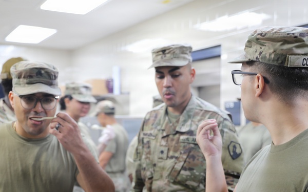 Military personnel in U.S. Army OCP (Operational Camouflage Pattern) uniforms are gathered in what appears to be a military kitchen or dining facility. Several soldiers are tasting or sampling food, with one soldier wearing a name tape reading 'COLON' visible on the right. The setting suggests a food preparation, quality control, or culinary training activity.