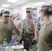 Military personnel in U.S. Army OCP (Operational Camouflage Pattern) uniforms are gathered in what appears to be a military kitchen or dining facility. Several soldiers are tasting or sampling food, with one soldier wearing a name tape reading 'COLON' visible on the right. The setting suggests a food preparation, quality control, or culinary training activity.
