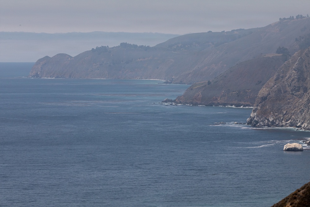 A coastal landscape photograph showing rugged cliffs and headlands meeting the ocean, likely along the California coast. The scene features layered promontories receding into a hazy distance, with calm blue-gray water in the foreground. The terrain is characterized by steep, rocky bluffs with sparse dry vegetation.