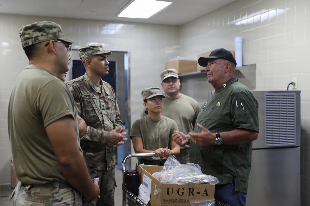 A group of military personnel in Army Combat Uniforms (ACU) are gathered in what appears to be a military kitchen or food service facility. An older civilian or senior official in a green shirt and black cap is addressing the soldiers, appearing to provide instruction or briefing. A UGR-A (Unitized Group Ration - A) box labeled 'Breakfast Menu 2' is visible on a cart in the foreground.