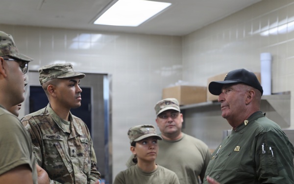 A group of military personnel in Army Combat Uniforms (ACU) are gathered in what appears to be a military kitchen or food service facility. An older civilian or senior official in a green shirt and black cap is addressing the soldiers, appearing to provide instruction or briefing. A UGR-A (Unitized Group Ration - A) box labeled 'Breakfast Menu 2' is visible on a cart in the foreground.