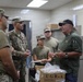 A group of military personnel in Army Combat Uniforms (ACU) are gathered in what appears to be a military kitchen or food service facility. An older civilian or senior official in a green shirt and black cap is addressing the soldiers, appearing to provide instruction or briefing. A UGR-A (Unitized Group Ration - A) box labeled 'Breakfast Menu 2' is visible on a cart in the foreground.