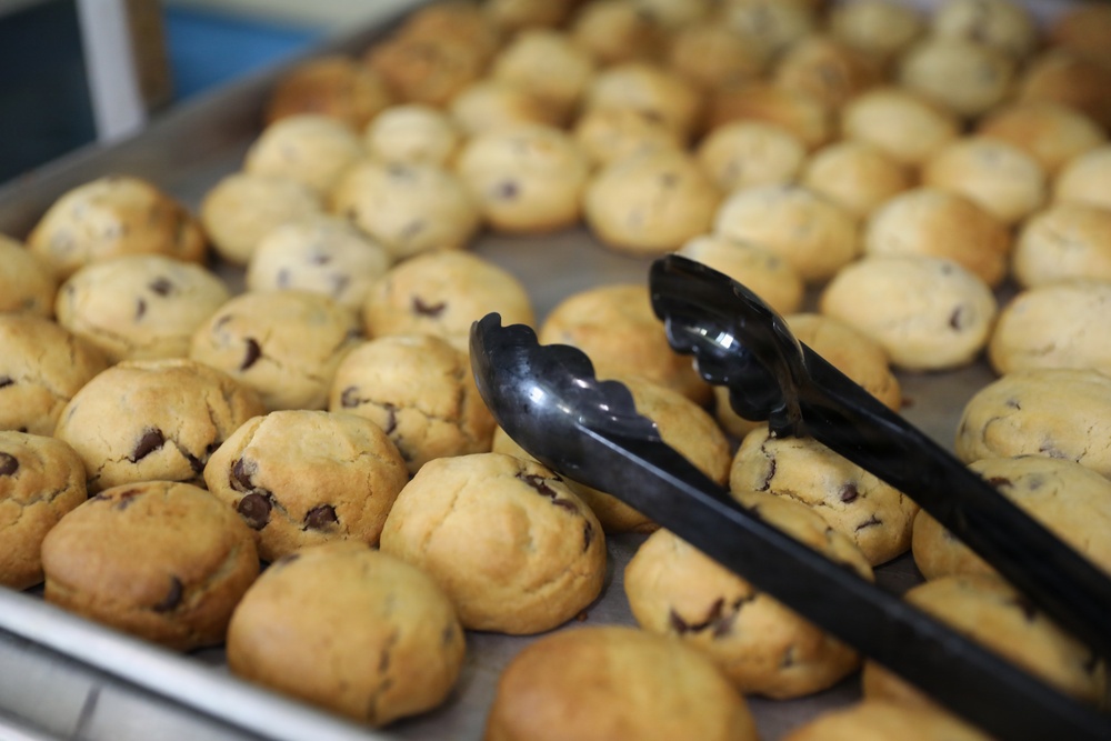 A large baking tray filled with freshly baked chocolate chip cookies arranged in rows. A pair of black plastic serving tongs rests on top of the cookies in the foreground. The setting appears to be a commercial or institutional kitchen or bakery.