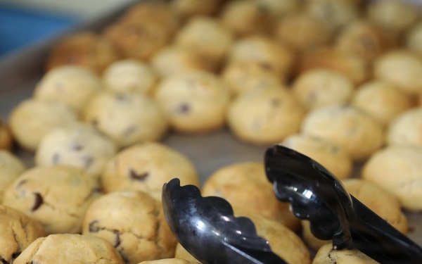 A large baking tray filled with freshly baked chocolate chip cookies arranged in rows. A pair of black plastic serving tongs rests on top of the cookies in the foreground. The setting appears to be a commercial or institutional kitchen or bakery.