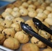 A large baking tray filled with freshly baked chocolate chip cookies arranged in rows. A pair of black plastic serving tongs rests on top of the cookies in the foreground. The setting appears to be a commercial or institutional kitchen or bakery.