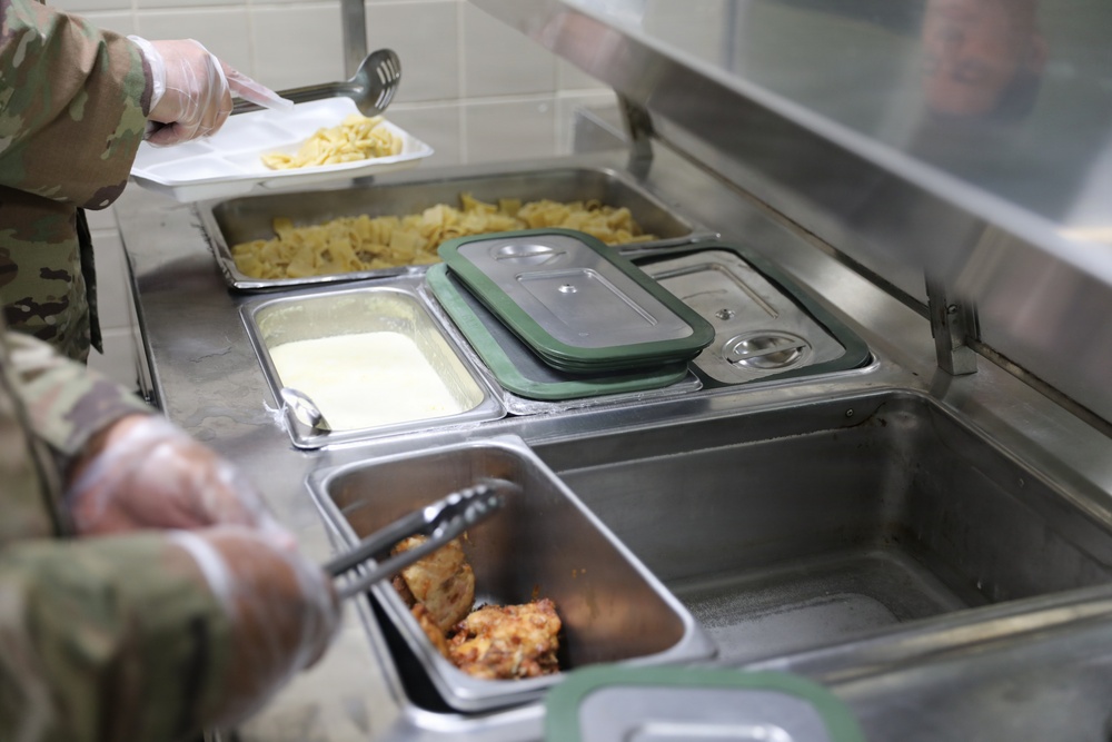 The image shows a military dining facility (DFAC) serving line where food is being portioned onto trays. Two individuals wearing camouflage uniforms and disposable plastic gloves are serving food from stainless steel hotel pans. The scene depicts an institutional food service environment with multiple food items being plated.