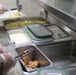 The image shows a military dining facility (DFAC) serving line where food is being portioned onto trays. Two individuals wearing camouflage uniforms and disposable plastic gloves are serving food from stainless steel hotel pans. The scene depicts an institutional food service environment with multiple food items being plated.
