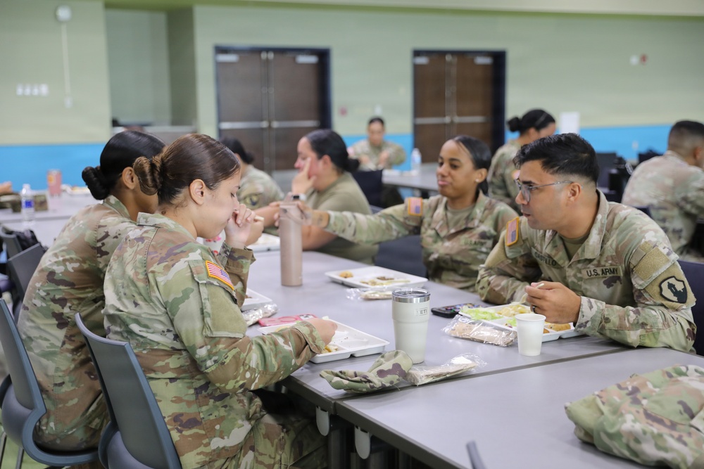 A group of U.S. Army soldiers in Operational Camouflage Pattern (OCP) uniforms are eating a meal together in what appears to be a military dining facility (DFAC). The soldiers are seated at gray cafeteria-style tables with food trays, beverages, and personal items visible. The setting is an institutional dining hall with green and blue painted walls.