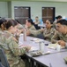 A group of U.S. Army soldiers in Operational Camouflage Pattern (OCP) uniforms are eating a meal together in what appears to be a military dining facility (DFAC). The soldiers are seated at gray cafeteria-style tables with food trays, beverages, and personal items visible. The setting is an institutional dining hall with green and blue painted walls.