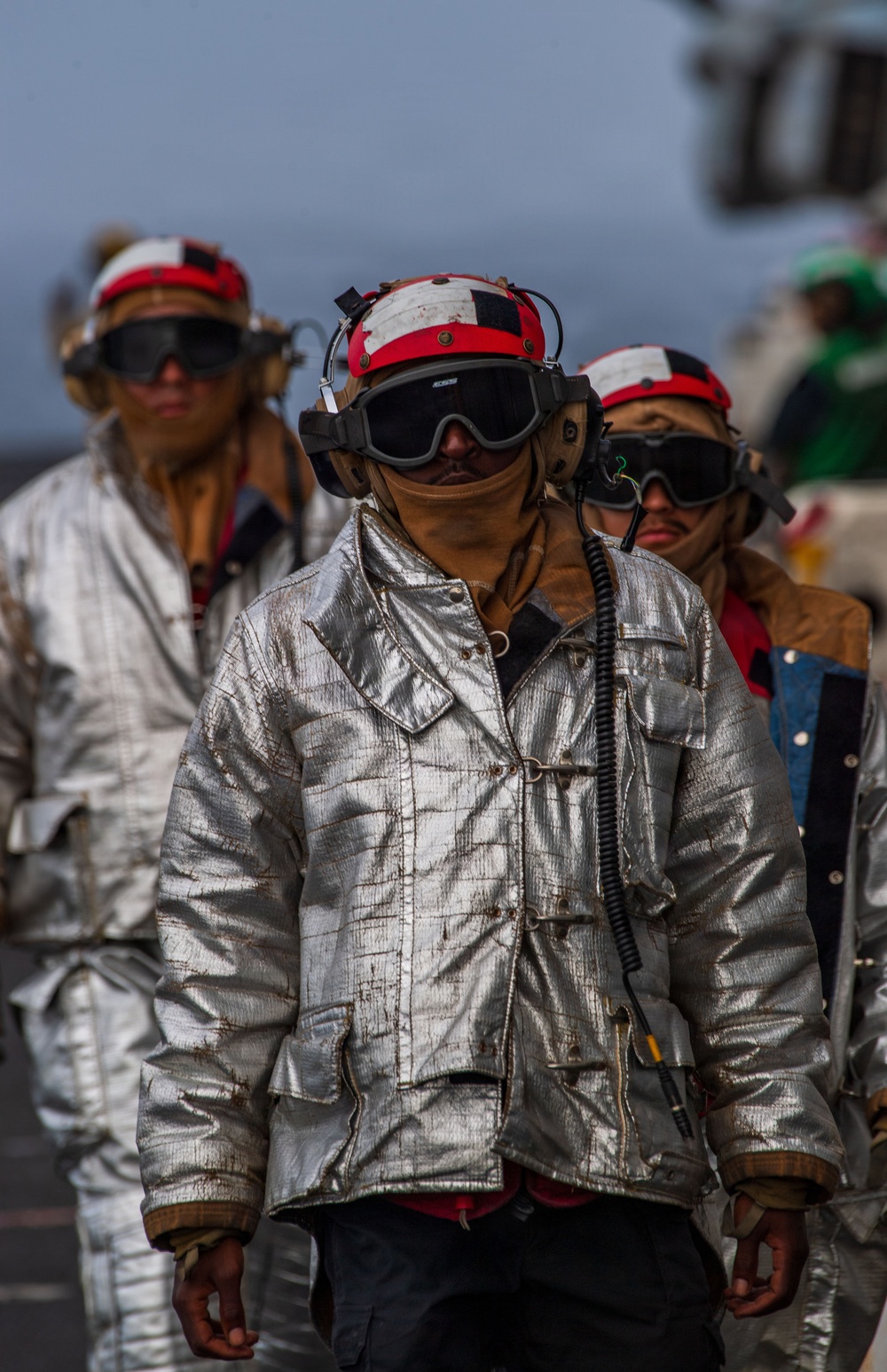 The image shows multiple personnel wearing aluminized proximity firefighting suits and red/white flight deck helmets with communication headsets and dark protective goggles. They appear to be walking in formation on what is likely a naval aircraft carrier flight deck. The gear is consistent with crash and salvage or firefighting crews assigned to carrier flight deck operations.