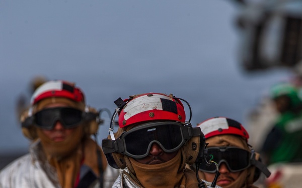 The image shows multiple personnel wearing aluminized proximity firefighting suits and red/white flight deck helmets with communication headsets and dark protective goggles. They appear to be walking in formation on what is likely a naval aircraft carrier flight deck. The gear is consistent with crash and salvage or firefighting crews assigned to carrier flight deck operations.
