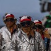 The image shows multiple personnel wearing aluminized proximity firefighting suits and red/white flight deck helmets with communication headsets and dark protective goggles. They appear to be walking in formation on what is likely a naval aircraft carrier flight deck. The gear is consistent with crash and salvage or firefighting crews assigned to carrier flight deck operations.