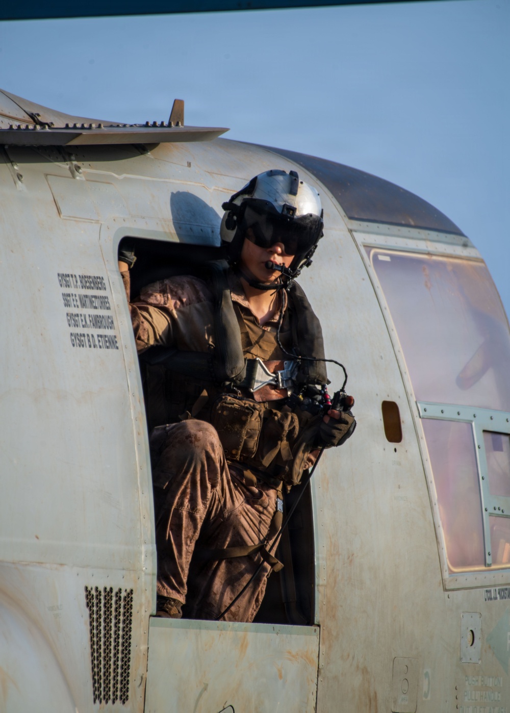 A military aircrew member is seated in the open door of a helicopter, wearing a flight helmet with visor and communications equipment. The individual is dressed in camouflage flight suit and tactical vest, holding what appears to be a control device or intercom unit. Text stenciled on the aircraft fuselage lists several names with the rank designation 'GYSGT' (Gunnery Sergeant), indicating a US Marine Corps aircraft.
