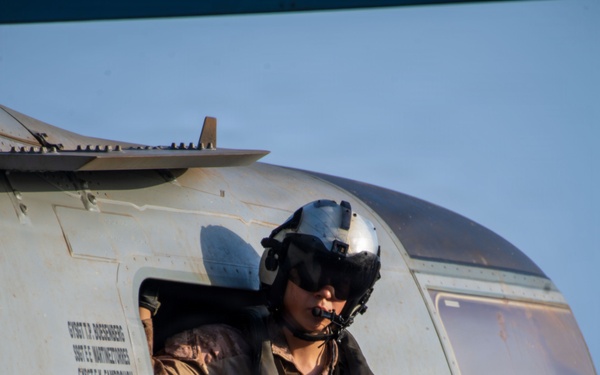 A military aircrew member is seated in the open door of a helicopter, wearing a flight helmet with visor and communications equipment. The individual is dressed in camouflage flight suit and tactical vest, holding what appears to be a control device or intercom unit. Text stenciled on the aircraft fuselage lists several names with the rank designation 'GYSGT' (Gunnery Sergeant), indicating a US Marine Corps aircraft.