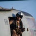 A military aircrew member is seated in the open door of a helicopter, wearing a flight helmet with visor and communications equipment. The individual is dressed in camouflage flight suit and tactical vest, holding what appears to be a control device or intercom unit. Text stenciled on the aircraft fuselage lists several names with the rank designation 'GYSGT' (Gunnery Sergeant), indicating a US Marine Corps aircraft.