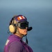 A U.S. Navy aviation fuels handler ('Grapes') stands on what appears to be an aircraft carrier flight deck, identifiable by the distinctive purple jersey and helmet. The individual is equipped with standard flight deck safety gear including hearing protection, ESS goggles, a communication headset, and black gloves. The ocean is visible in the background, confirming an at-sea operational environment.
