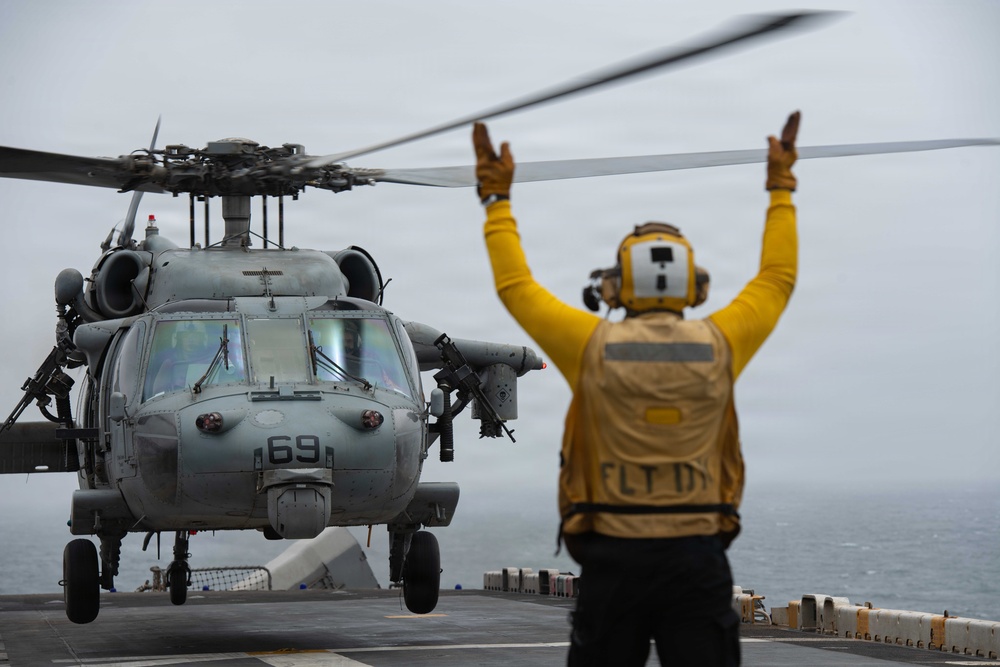 A naval flight deck director in yellow gear is signaling with both arms raised to guide a military helicopter numbered '69' during flight deck operations aboard a naval vessel at sea. The helicopter appears to be an MH-60 Seahawk in gray military livery, hovering or preparing to land on the ship's flight deck. The overcast sky and open ocean are visible in the background.