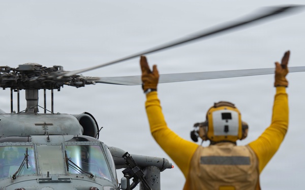 A naval flight deck director in yellow gear is signaling with both arms raised to guide a military helicopter numbered '69' during flight deck operations aboard a naval vessel at sea. The helicopter appears to be an MH-60 Seahawk in gray military livery, hovering or preparing to land on the ship's flight deck. The overcast sky and open ocean are visible in the background.