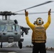 A naval flight deck director in yellow gear is signaling with both arms raised to guide a military helicopter numbered '69' during flight deck operations aboard a naval vessel at sea. The helicopter appears to be an MH-60 Seahawk in gray military livery, hovering or preparing to land on the ship's flight deck. The overcast sky and open ocean are visible in the background.