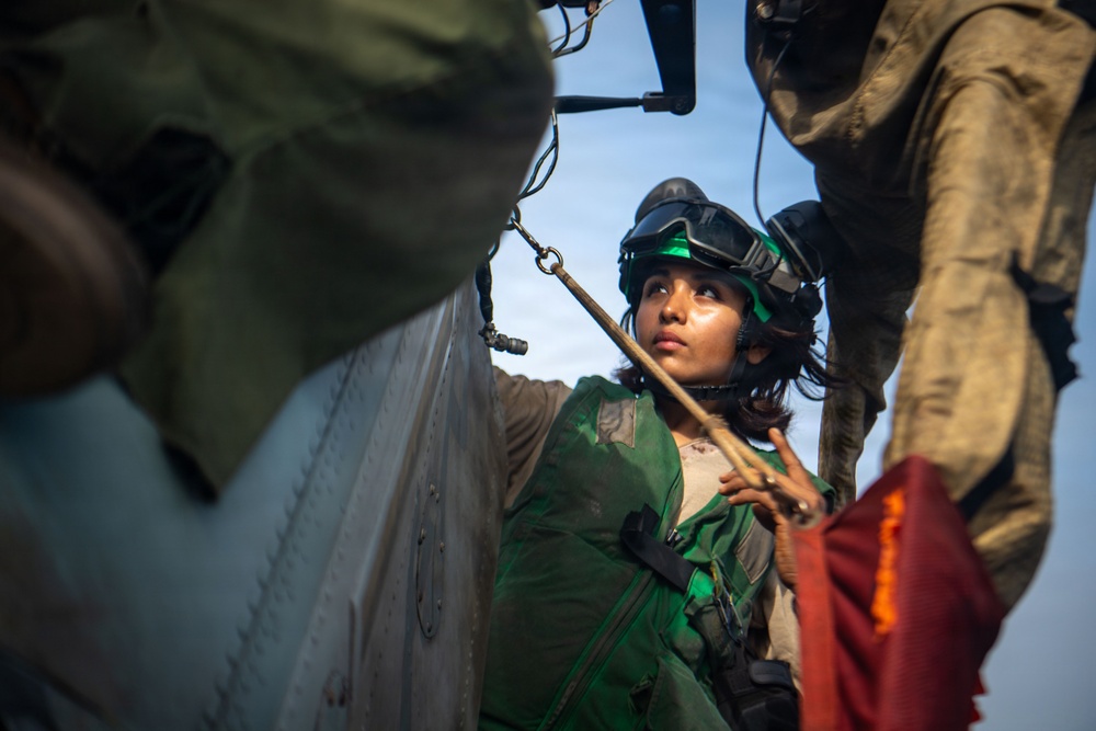 A military service member wearing a green flight suit and helmet with goggles is working near the exterior of an aircraft, likely a helicopter. The individual appears to be performing pre-flight or maintenance checks, handling a tether or safety line. The image is taken from a low, close-up angle with other personnel partially visible in the frame.