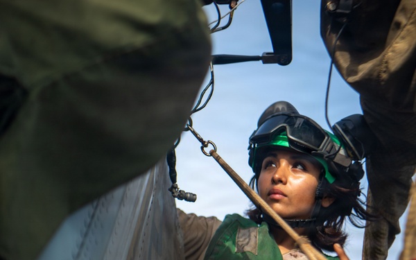 A military service member wearing a green flight suit and helmet with goggles is working near the exterior of an aircraft, likely a helicopter. The individual appears to be performing pre-flight or maintenance checks, handling a tether or safety line. The image is taken from a low, close-up angle with other personnel partially visible in the frame.