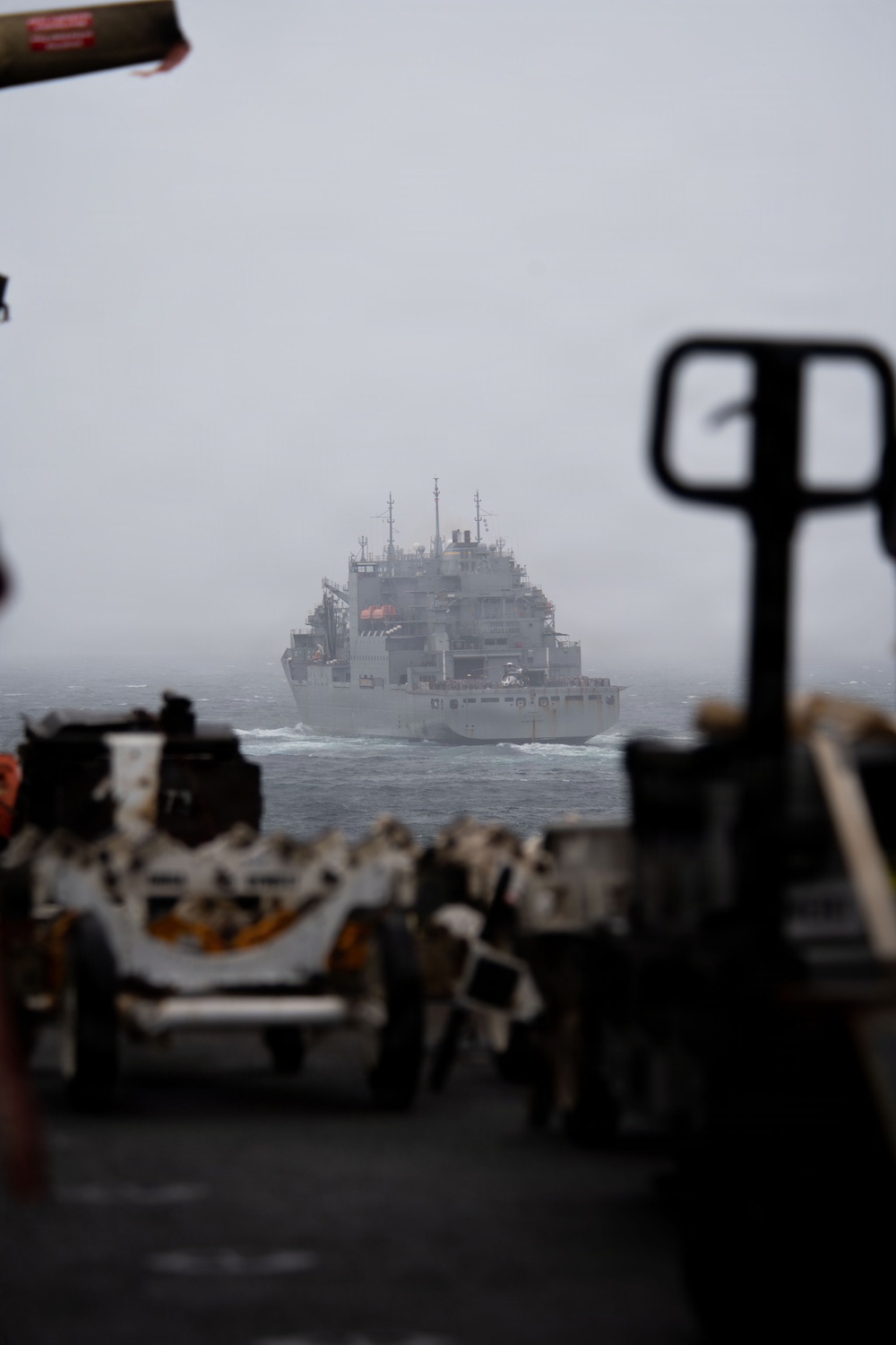 A large gray naval auxiliary or replenishment vessel is visible at sea through fog or haze, photographed from the deck of another ship. The foreground shows out-of-focus deck equipment, machinery, and what appears to be a pallet jack or deck handling equipment. Conditions appear overcast and misty, suggesting open ocean operations.