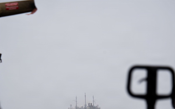 A large gray naval auxiliary or replenishment vessel is visible at sea through fog or haze, photographed from the deck of another ship. The foreground shows out-of-focus deck equipment, machinery, and what appears to be a pallet jack or deck handling equipment. Conditions appear overcast and misty, suggesting open ocean operations.