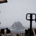 A large gray naval auxiliary or replenishment vessel is visible at sea through fog or haze, photographed from the deck of another ship. The foreground shows out-of-focus deck equipment, machinery, and what appears to be a pallet jack or deck handling equipment. Conditions appear overcast and misty, suggesting open ocean operations.