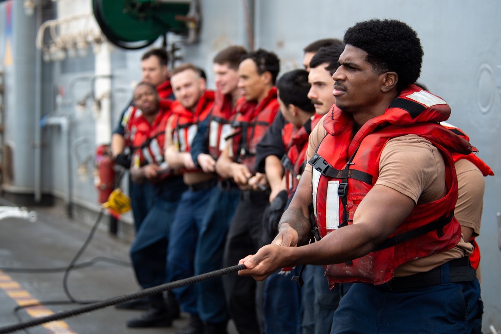 A group of naval personnel wearing red life vests and dark blue uniforms are lined up on the deck of a military vessel, collectively pulling a thick rope in a coordinated effort. The scene appears to depict a seamanship or line-handling evolution, likely during a replenishment-at-sea or mooring operation. The foreground sailor is in sharp focus while the line of crew members extends into the background.