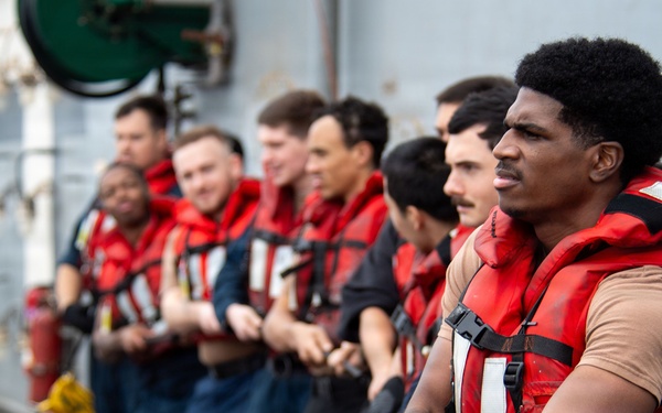 A group of naval personnel wearing red life vests and dark blue uniforms are lined up on the deck of a military vessel, collectively pulling a thick rope in a coordinated effort. The scene appears to depict a seamanship or line-handling evolution, likely during a replenishment-at-sea or mooring operation. The foreground sailor is in sharp focus while the line of crew members extends into the background.