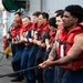 A group of naval personnel wearing red life vests and dark blue uniforms are lined up on the deck of a military vessel, collectively pulling a thick rope in a coordinated effort. The scene appears to depict a seamanship or line-handling evolution, likely during a replenishment-at-sea or mooring operation. The foreground sailor is in sharp focus while the line of crew members extends into the background.