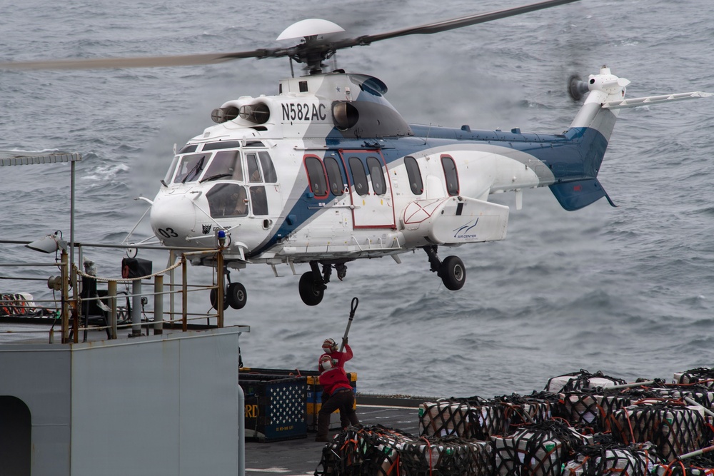 A white and blue civilian helicopter with registration N582AC is hovering over the deck of a naval or military vessel at sea. A deck crew member in red and safety gear is visible below the helicopter, appearing to guide or signal the aircraft. The deck is loaded with netted cargo pallets, suggesting a vertical replenishment (VERTREP) or logistics operation.