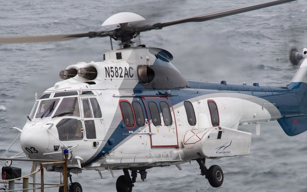 A white and blue civilian helicopter with registration N582AC is hovering over the deck of a naval or military vessel at sea. A deck crew member in red and safety gear is visible below the helicopter, appearing to guide or signal the aircraft. The deck is loaded with netted cargo pallets, suggesting a vertical replenishment (VERTREP) or logistics operation.