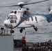 A white and blue civilian helicopter with registration N582AC is hovering over the deck of a naval or military vessel at sea. A deck crew member in red and safety gear is visible below the helicopter, appearing to guide or signal the aircraft. The deck is loaded with netted cargo pallets, suggesting a vertical replenishment (VERTREP) or logistics operation.