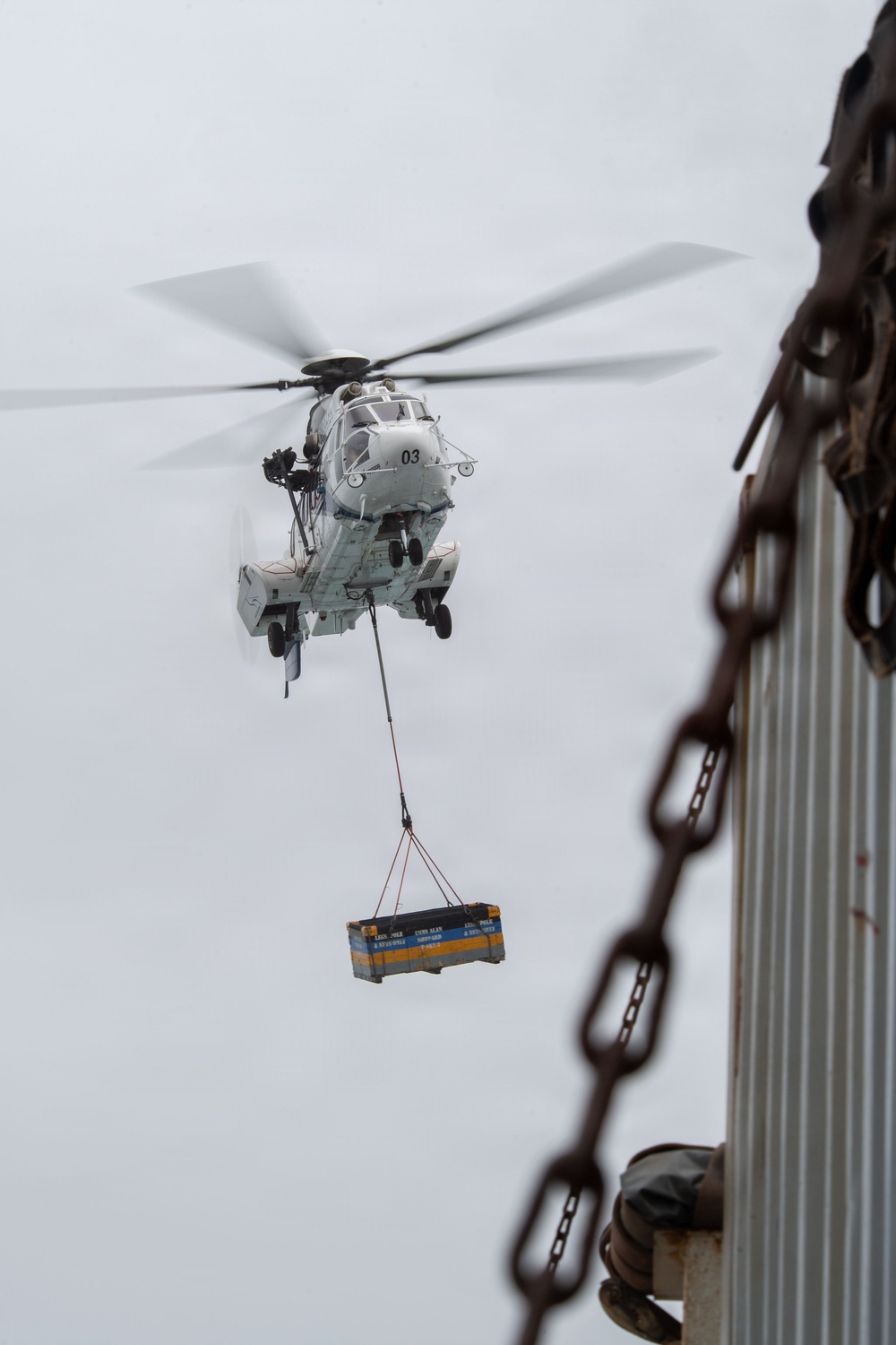 A military helicopter marked with the number '03' is performing a sling-load cargo operation, carrying a blue and yellow labeled container suspended by cables beneath its fuselage. The image is taken from an elevated vantage point near a corrugated metal structure with a large rusty chain in the foreground. The overcast sky provides a neutral background, emphasizing the helicopter and its suspended load.