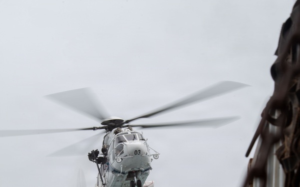 A military helicopter marked with the number '03' is performing a sling-load cargo operation, carrying a blue and yellow labeled container suspended by cables beneath its fuselage. The image is taken from an elevated vantage point near a corrugated metal structure with a large rusty chain in the foreground. The overcast sky provides a neutral background, emphasizing the helicopter and its suspended load.