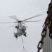 A military helicopter marked with the number '03' is performing a sling-load cargo operation, carrying a blue and yellow labeled container suspended by cables beneath its fuselage. The image is taken from an elevated vantage point near a corrugated metal structure with a large rusty chain in the foreground. The overcast sky provides a neutral background, emphasizing the helicopter and its suspended load.