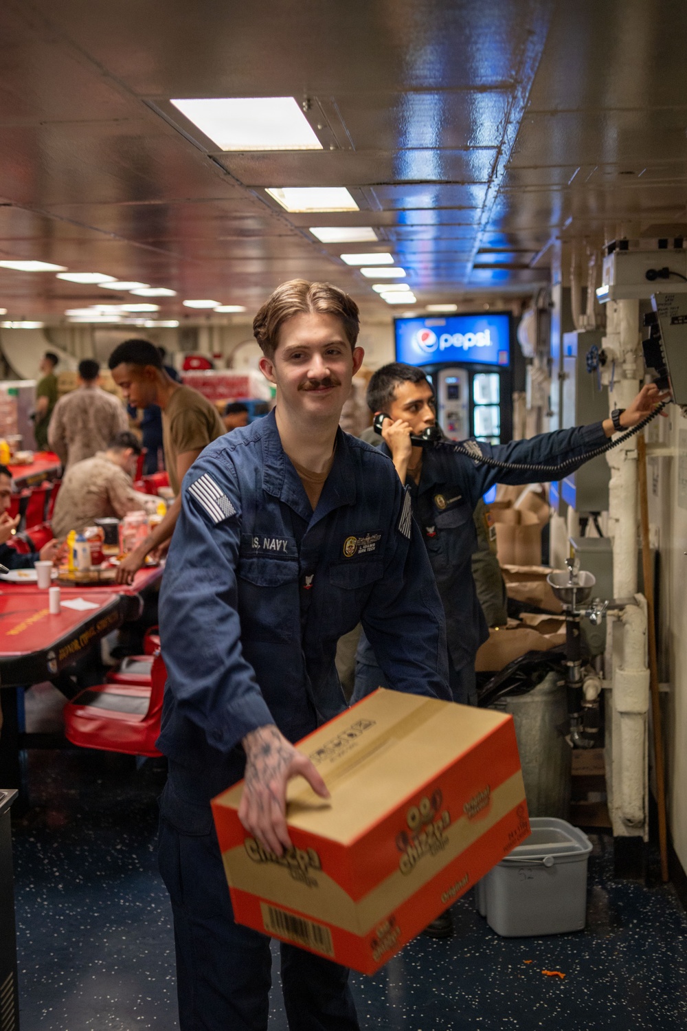 A U.S. Navy sailor in blue coveralls smiles at the camera while carrying a cardboard box in what appears to be a ship's mess hall or galley. Behind him, another sailor in dark coveralls is using a corded telephone mounted to the wall. The setting is a naval vessel interior with red dining tables, fluorescent lighting, and a Pepsi vending machine visible in the background.