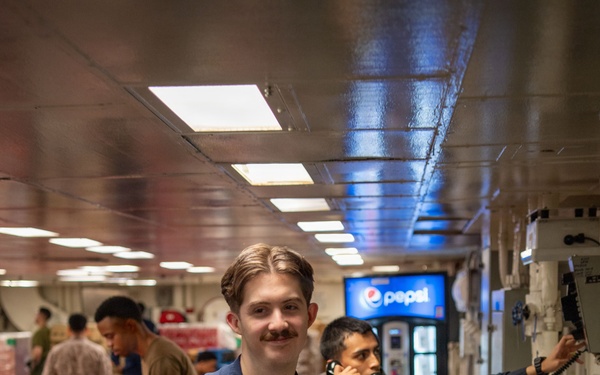 A U.S. Navy sailor in blue coveralls smiles at the camera while carrying a cardboard box in what appears to be a ship's mess hall or galley. Behind him, another sailor in dark coveralls is using a corded telephone mounted to the wall. The setting is a naval vessel interior with red dining tables, fluorescent lighting, and a Pepsi vending machine visible in the background.