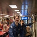 A U.S. Navy sailor in blue coveralls smiles at the camera while carrying a cardboard box in what appears to be a ship's mess hall or galley. Behind him, another sailor in dark coveralls is using a corded telephone mounted to the wall. The setting is a naval vessel interior with red dining tables, fluorescent lighting, and a Pepsi vending machine visible in the background.