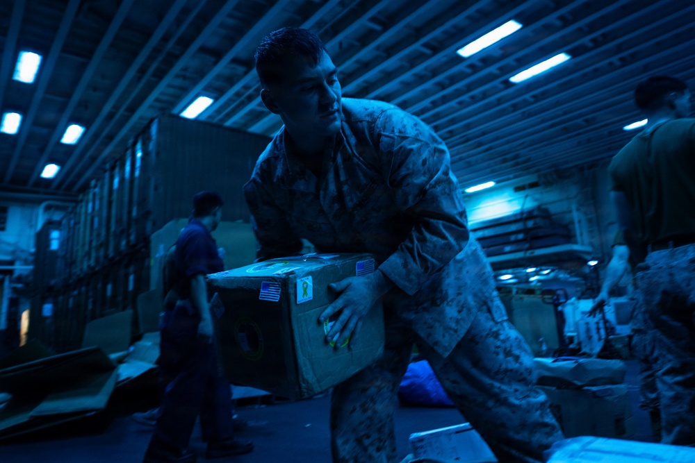 Military personnel in camouflage uniforms are handling and moving cardboard boxes in what appears to be the interior cargo hold or hangar deck of a naval vessel. The scene is illuminated by blue-tinted fluorescent lighting, creating a dim, industrial atmosphere. The central figure is actively carrying a large cardboard box marked with a US flag sticker and a yellow ribbon sticker.