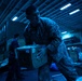 Military personnel in camouflage uniforms are handling and moving cardboard boxes in what appears to be the interior cargo hold or hangar deck of a naval vessel. The scene is illuminated by blue-tinted fluorescent lighting, creating a dim, industrial atmosphere. The central figure is actively carrying a large cardboard box marked with a US flag sticker and a yellow ribbon sticker.