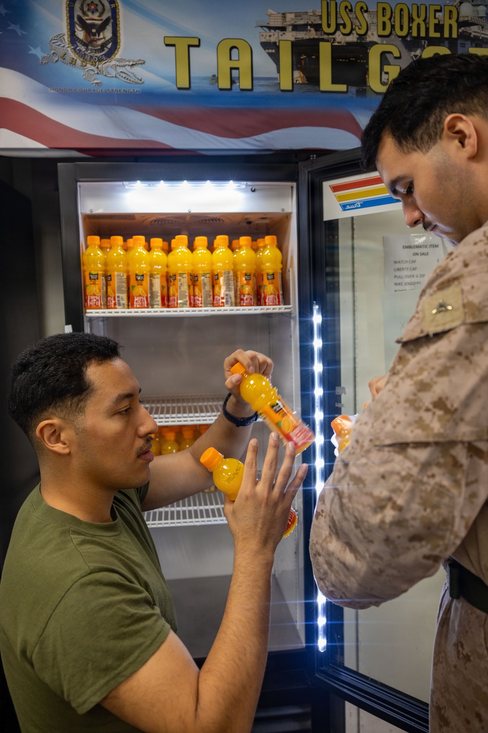Two military personnel are interacting near a refrigerator stocked with orange juice bottles at a USS Boxer Tailgate event. One individual is wearing a green t-shirt and the other is in Marine Corps MARPAT camouflage uniform. They appear to be exchanging or examining orange juice bottles together.