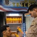 Two military personnel are interacting near a refrigerator stocked with orange juice bottles at a USS Boxer Tailgate event. One individual is wearing a green t-shirt and the other is in Marine Corps MARPAT camouflage uniform. They appear to be exchanging or examining orange juice bottles together.