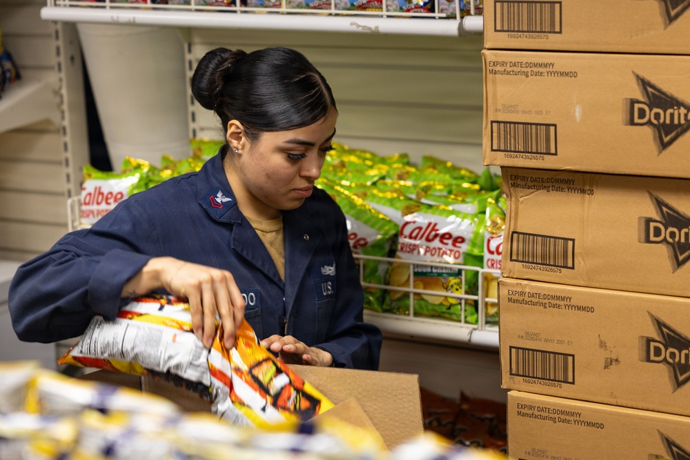 A U.S. military service member in navy blue utility uniform is stocking or organizing snack food items in what appears to be a military exchange or commissary store. She is handling chip bags near stacked cardboard boxes of Doritos, with Calbee Crispy Potato chip bags visible on a wire shelf behind her. The setting suggests a retail or supply storage area on a military installation.